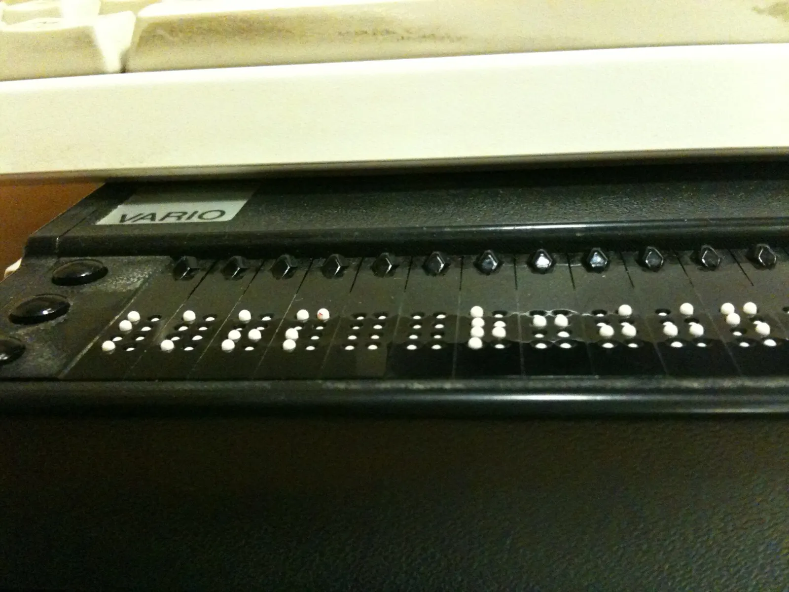 A refreshable braille display: a long, low device with a row of cells, each raising small rounded pins to form braille characters, and round control buttons along the front edge.